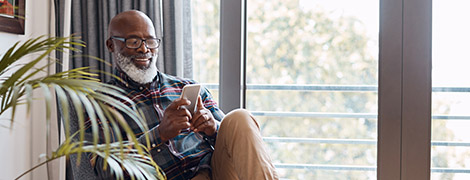 A elderly man with a beard sitting in a comfortable chair by a window and downloading the MyChart app on his smartphone.