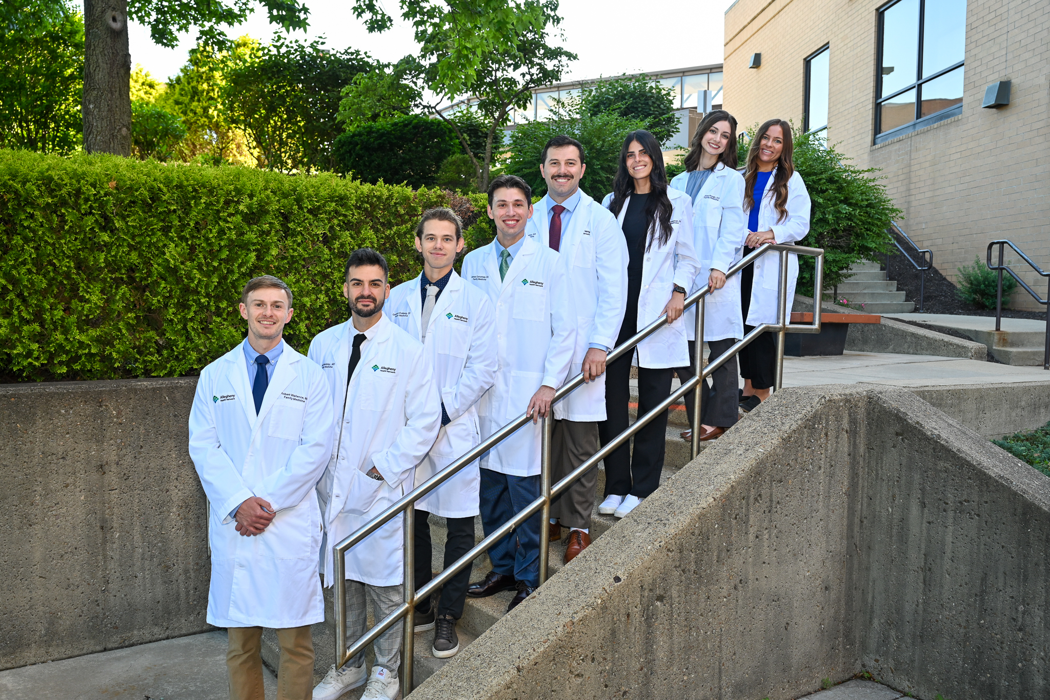 Eight Allegheny Health Network medical residents, 5 men and 3 women, smiling in white lab coats on an outdoor staircase with lush greenery and a building in the background.