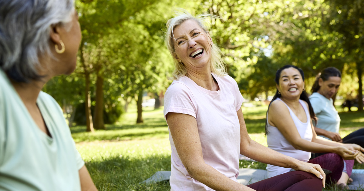 Mature female is laughing while enjoying yoga with female friends in a public park.
