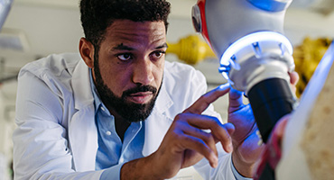A black, male medical professional in a lab coat carefully examines a robotic arm with a blue light.