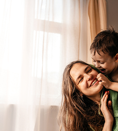 A small boy climbing on his mother's back while she smiles looking back at him