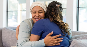 A female patient happily hugs a female healthcare worker on a couch.