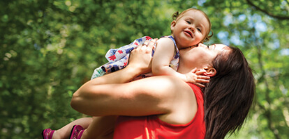 A woman giving her smiling toddler a kiss on the cheek as she holds her up in front of a grove of trees on a sunny summer day 