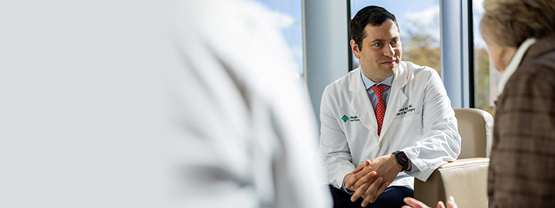 Doctor in white coat with red tie talking to an older female patient from across a room.