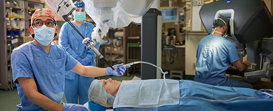 Surgeon with robotic system with a patient on table., as another professional at controls.