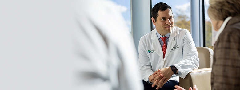 A white medical professional in a white coat talks to an older woman in a brown jacket, who smiles.