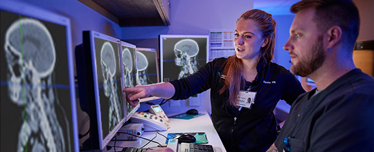 Two medical professionals in dark clothing look at brain scans on computer monitors.