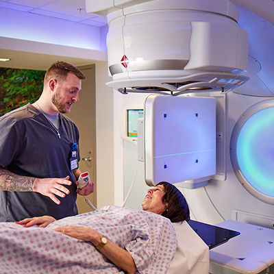 A medical professional in blue scrubs talks to a woman lying under a large machine.