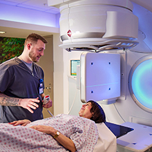 A medical professional in blue scrubs talks to a woman lying under a large machine.