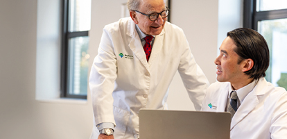 Two male medical professionals, one standing and one sitting, are looking at a laptop together.