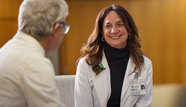 A medical professional wearing a white coat and name tag looks happy talking with someone.