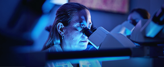 A woman scientist with long hair peers into a microscope in a blue-lit room.