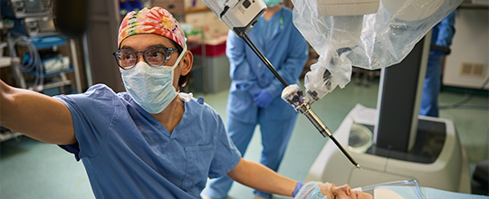 A medical professional in blue scrubs and a colorful cap oversees a robot near a patient.
