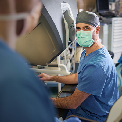 A white medical professional in blue scrubs, a gray cap, and a mask smiles while looking at the camera.
