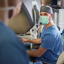 A white medical professional in blue scrubs, a gray cap, and a mask smiles while looking at the camera.