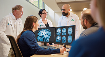 A group of medical professionals look at brain scans on two computer monitors in an office.