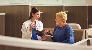 A medical professional in a white coat talks to an older woman in a waiting room.