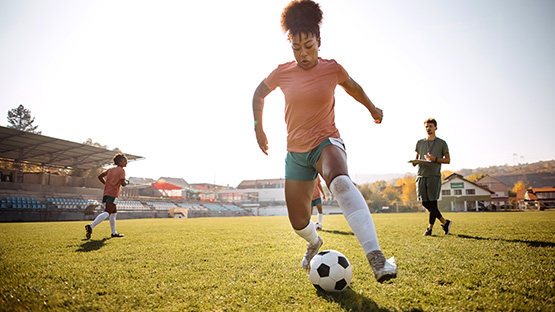 A woman in a pink shirt and green shorts dribbles a soccer ball on a sunny field during practice.