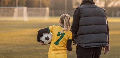 A parent holding hands with their child and walking across a soccer field.