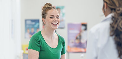 A smiling woman in a green shirt talks to a medical professional in a white coat.